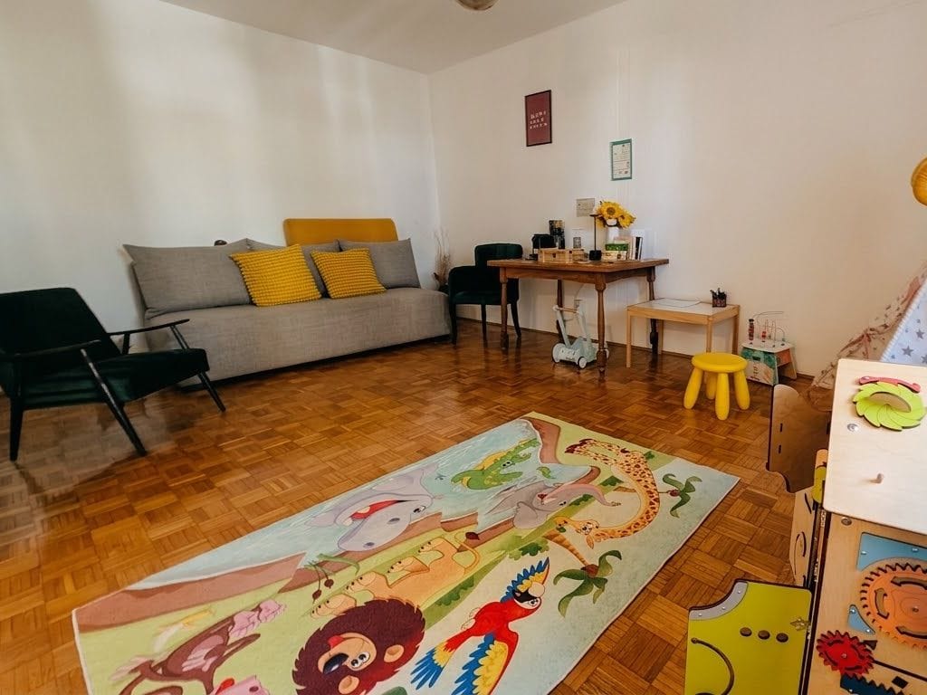 Čekaonica u Govorlendu: siva sofa sa žutim jastucima, zelena fotelja, sto sa kafom i suncokretima i šareni tepih sa životinjama