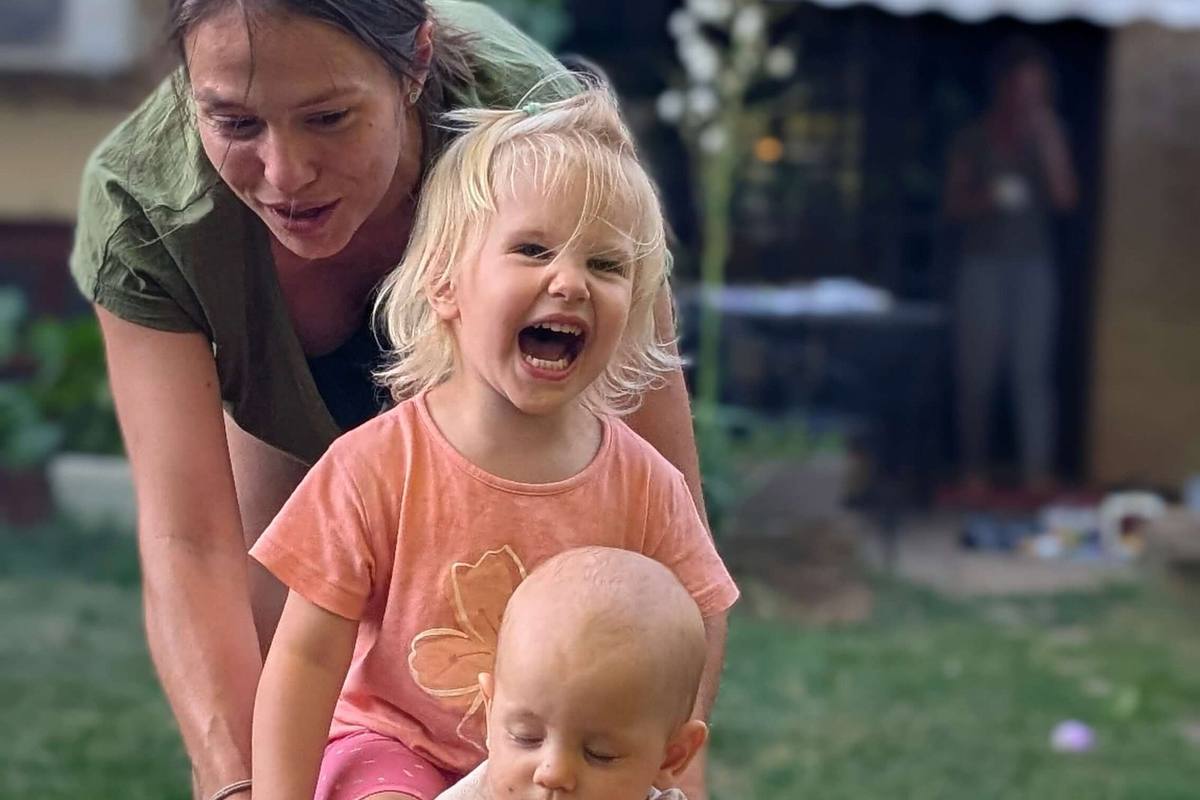 Sara gura dvoje dece u žutim baštenskim kolicima kroz dvorište, a pas trči pored njih