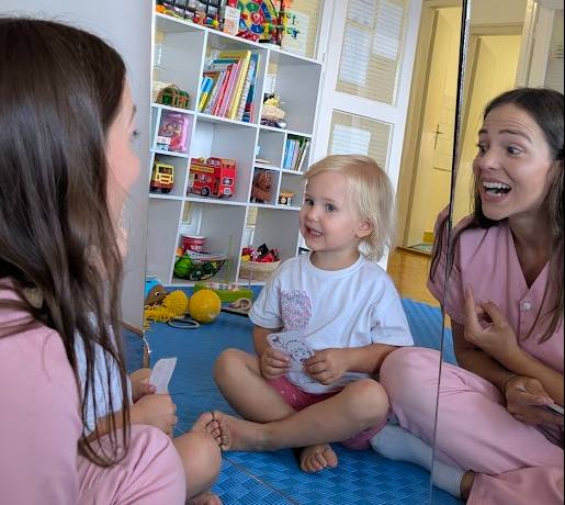 Speech Language Pathologist and child sit on a blue mat in a colorful playroom with shelves. The woman smiles and talks, creating a joyful atmosphere.
